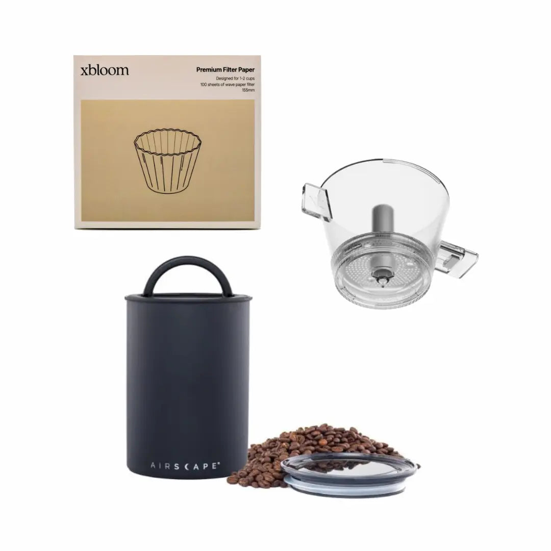 Collection of coffee brewing equipment including a box of x bloom filter paper, a black airscape container, a glass coffee grinder, and coffee beans.