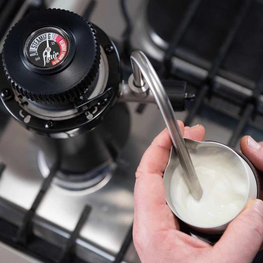 hands holding a container of milk with a steam wand texturing the milk for a coffee, Flair Wizard milk steamer in the background Basic Barista Australia
