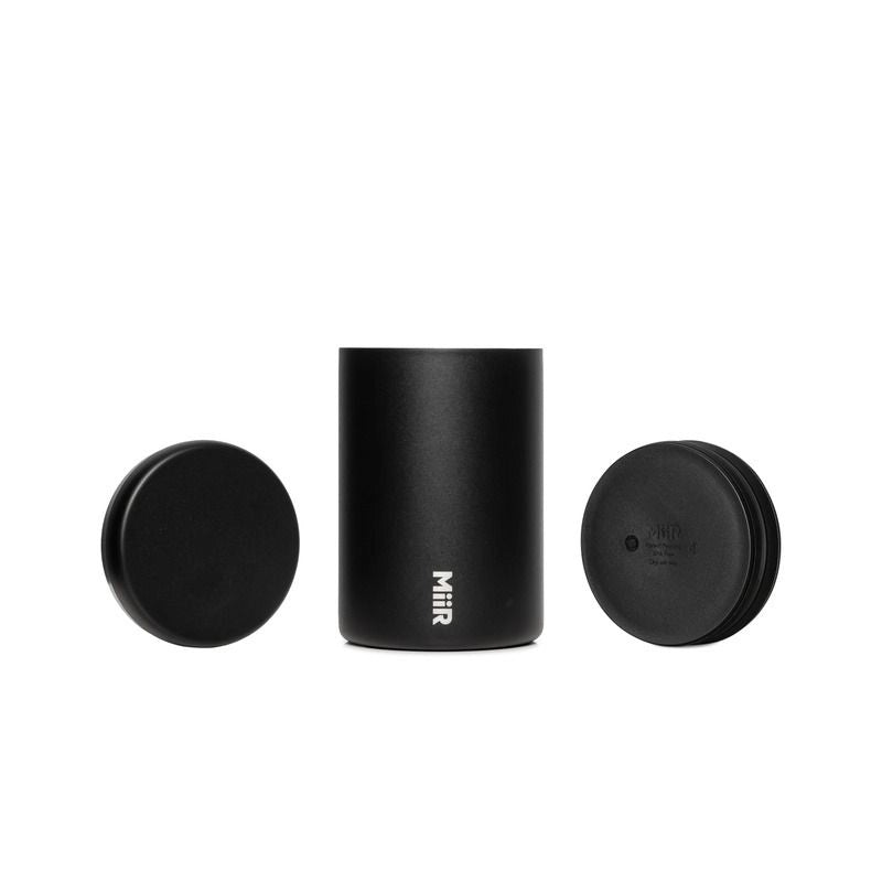 A matte black cylindrical insulated container with a matching lid and insert plunger, featuring the vertical white 'Miir' logo near the bottom, set against a plain white background.
Miir Airlock Coffee Storage Container