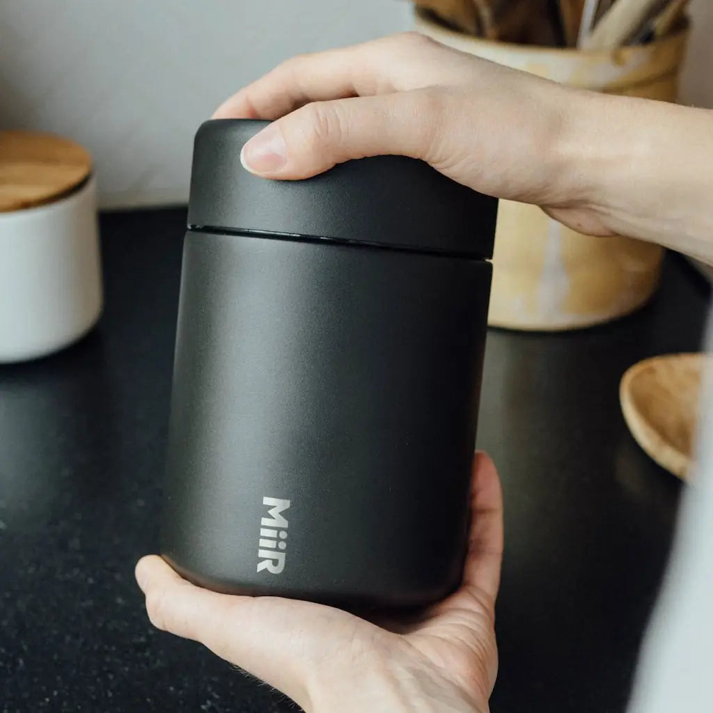 Person holding a black MiiR insulated coffee storage container on a kitchen counter.