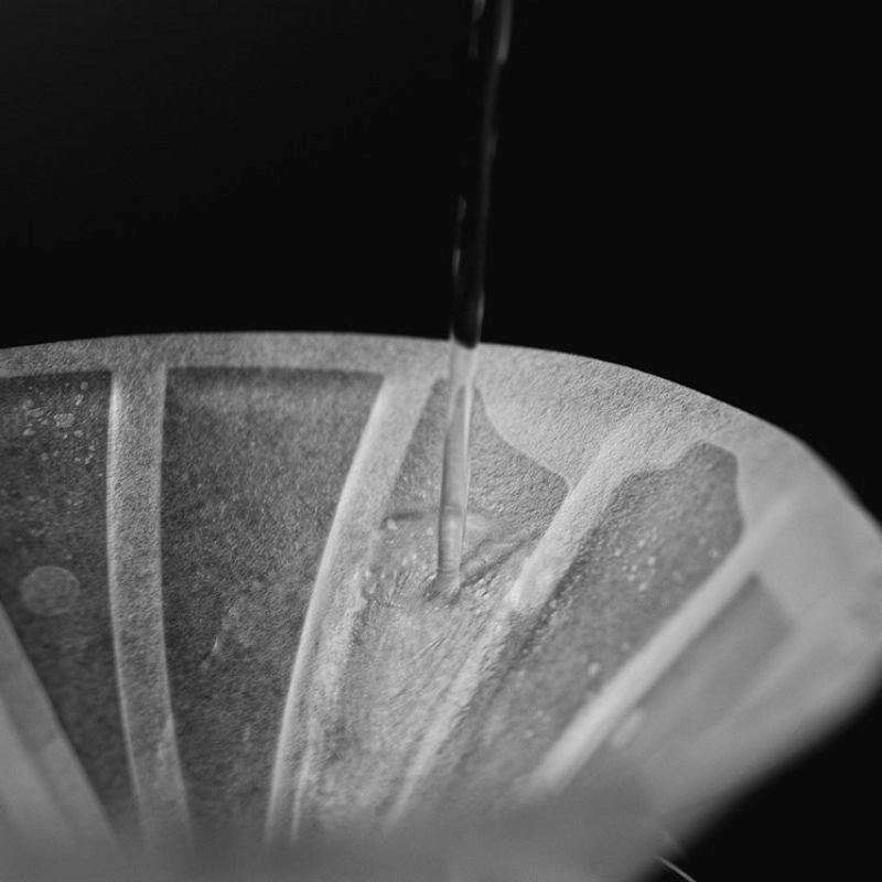 Black and white image of a UFO Dripper  with a slow pour of hot water being poured into the Sibarist UFO Dripper filter collaboration - Black Background Basic Barista Photography