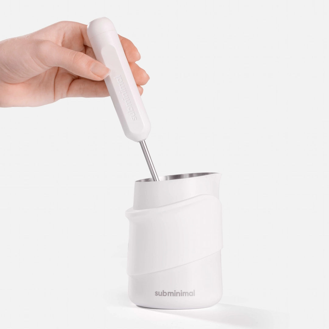 A hand holding a white electric milk frother above a white Subminimal insulated container with a stainless steel insert, set against a clean white background.